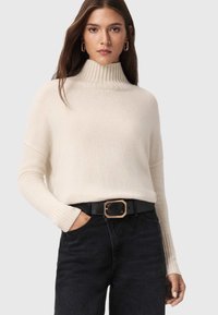 Kremowy sweter z dzianiny z wysokim prążkowanym kołnierzem. W połączeniu z czarnymi dżinsami i szerokim czarnym paskiem z złotą klamrą.