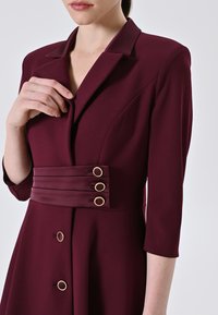 Bordeauxfarbiges Kleid mit Kragen, Dreiviertelärmeln und tailliertem Schnitt, der drei goldene Ringschmuckakzente und eine glatte Textur aufweist.