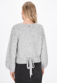 Femeie cu păr blond poartă un pulover tricotat de culoare gri deschis, cu mâneci late și se leagă la partea inferioară a spatelui, asortat cu blugi negri.
