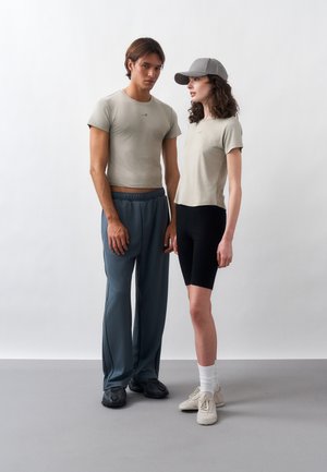 Mann og kvinne står side om side, begge iført lysegrå t-skjorter; mannen i løse blå bukser og joggesko, kvinnen i svarte shorts, caps, sokker og joggesko.