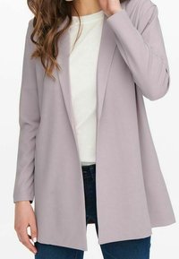 Blazer grigio chiaro realizzato in tessuto liscio e elasticizzato, con colletto a rever e design aperto, abbinato a una parte superiore bianca e jeans blu.