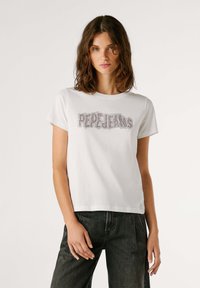 Camiseta blanca de algodón con mangas cortas que presenta "PERLEJANS" en adornos de cristales en el pecho. Combinada con jeans negros.