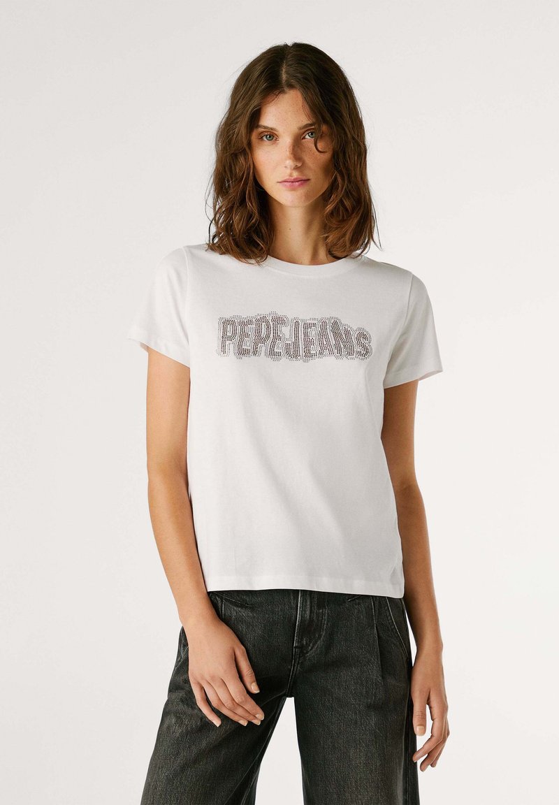 Camiseta blanca de algodón con mangas cortas que presenta "PERLEJANS" en adornos de cristales en el pecho. Combinada con jeans negros.