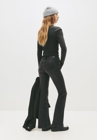 Kvinna som bär grå mössa, svart långärmad topp, mörka utsvängda jeans, håller en svart jacka i ena handen, står mot en enkel vit bakgrund.