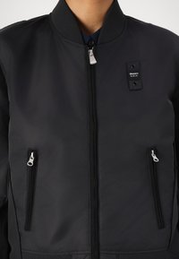 Čierna bomber bunda so zipsom na prednej strane, rebrovaným golierom a dvoma bočnými vreckami so zipsom. Obsahuje malú nášivku loga na ľavom hrudníku.