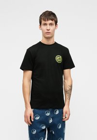 Νεαρός άνδρας με κοντά καστανά μαλλιά, φορώντας μια μαύρη μπλούζα Santa Cruz και μπλε τζιν τυπωμένα με λευκά σύμβολα γιν-γιανγκ, που στέκεται σε λευκό φόντο.