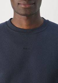 Gros plan d'une personne portant un sweat-shirt bleu marine à col rond avec le texte "SANDRO" subtilement brodé sur la poitrine.