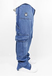 Blaue Denim-Cargo-Hosen mit mehreren Taschen, versehen mit besticktem Kronsymbol. Lockerer Schnitt mit schmal zulaufenden Bündchen, kombiniert mit weißen Sneakers.