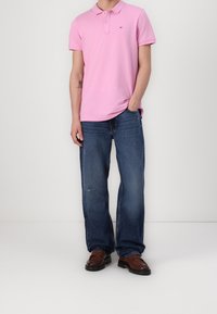 Polo shirt rose clair avec un col et un petit logo, associé à un jean en denim bleu ample et des chaussures brunes à enfiler. Design et coupe simples.