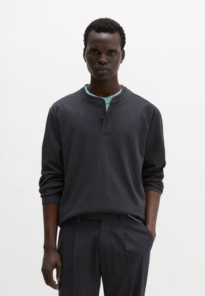 Mężczyzna stojący na jednolitym białym tle, ubrany w ciemną koszulę henley z długim rękawem na jasnoniebieskim podkoszulku i ciemne spodnie, z jedną ręką w kieszeni.