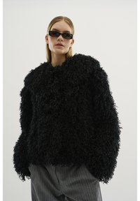Vrouw in een zwarte, krullende, oversized trui, met grijze krijtstreepbroek en slanke zwarte zonnebril, tegen een witte achtergrond.