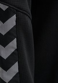 Nærbillede af sort stof med lodrette ribber og hvide chevron-striber, der er arrangeret lodret langs en søm.