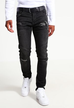 Jean noir délavé avec des panneaux côtelés aux genoux, porté avec une ceinture noire, une chemise blanche à manches longues et des baskets blanches.