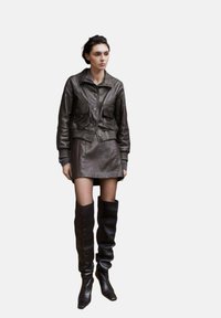 Jachetă scurtă din piele maro, cu buzunare frontale, asortată cu o fustă din piele și cizme până la genunchi. Design elegant și mulat.