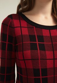 Röd och svart rutad tröja med en ribbad svart halsringning. Tyget har ett strukturerat mönster med växlande rutor.