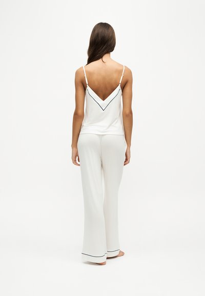 Femme debout pieds nus, de dos, portant un caraco blanc avec bordure noire et un pantalon large assorti sur fond uni.