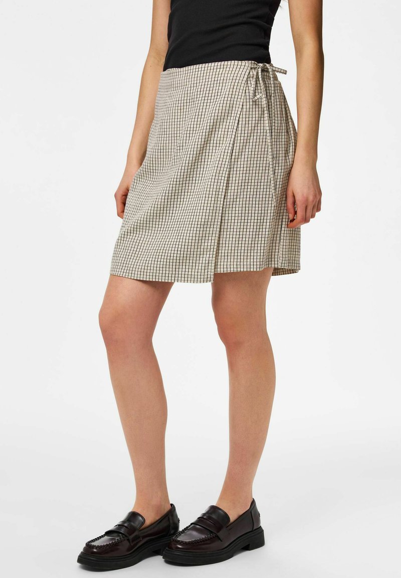 Femme portant une jupe portefeuille beige à motif quadrillé noir, un haut noir et des mocassins en cuir marron foncé, debout devant un fond blanc.