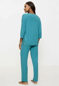 Set di abbigliamento loungewear color teal composto da una maglia ampia con scollo rotondo e maniche a tre quarti, abbinata a pantaloni a gamba dritta. Texture liscia.