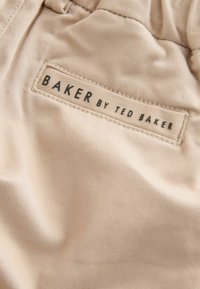 Primo piano di tessuto beige con etichetta rettangolare cucita con la scritta "BAKER BY TED BAKER" in testo nero.