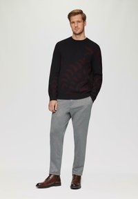 Zwart sweater met een bordeauxrood abstract patroon, gecombineerd met lichtgrijze broek en bruine leren laarzen, geplaatst tegen een neutrale achtergrond.