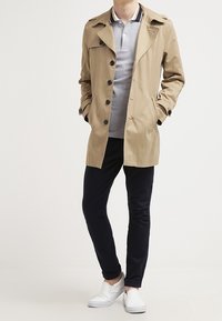 Beżowy płaszcz trenczowy z zapięciem na guziki, dużym kołnierzem i dwiema przednimi kieszeniami, noszony na szarym polo. Czarny dopasowany zestaw spodni i białe buty wsuwane.