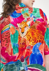 Kvinna som bär en färgglad skjorta med illustrationer av en leopard, tukan, toucanet, sengångare, blå fågel och tapir i livfulla röda, blå, orange och gröna nyanser.