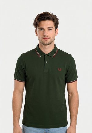 Man draagt een donkergroen poloshirt met rood-witte bies op de kraag en mouwen, staand tegen een effen witte achtergrond.