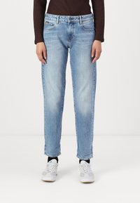 Osoba u svijetloplavim ravnim trapericama, tamnosmeđoj dugoj majici, crnim čarapama i bijelim tenisicama stoji naslonjena na jednostavnu pozadinu.