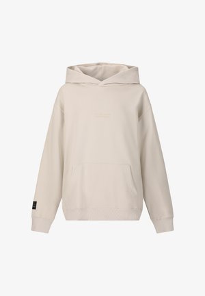 Jonge man staand met handen in de zakken, gekleed in een beige hoodie, zwarte slim-fit broek en zwarte sneakers, tegen een effen witte achtergrond.