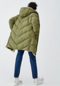 Abrigo acolchado verde oliva con capucha, con un diseño en forma de chevron, cremalleras laterales y forro suave. Combinado con vaqueros y zapatillas blancas.