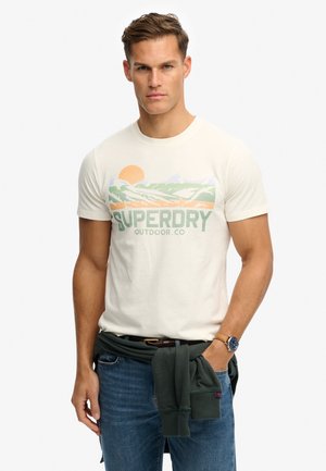 Mand iført en hvid Superdry t-shirt med landskabsprint, blå jeans og en mørkegrøn sweatshirt bundet omkring hans talje.