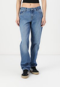 Jeans de mezclilla azul de cintura alta con un corte holgado, cinco bolsillos y costuras visibles. Combinados con zapatillas negras con rayas blancas.