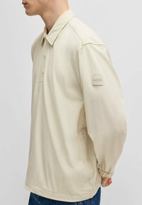 Helle beige Pulloverjacke mit Kragen, drei Knöpfen und elastischen Bündchen. Ausgestattet mit einem kleinen Stoff-Logopatch an den linken Ärmel.