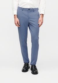 Μπλε παντελόνια ραμμένα από υφασμάτινη υφή, με εφαρμοστή σχεδίαση, πλαϊνές τσέπες και κλείσιμο με κουμπί. Φορεμένα με μαύρα δερμάτινα παπούτσια.