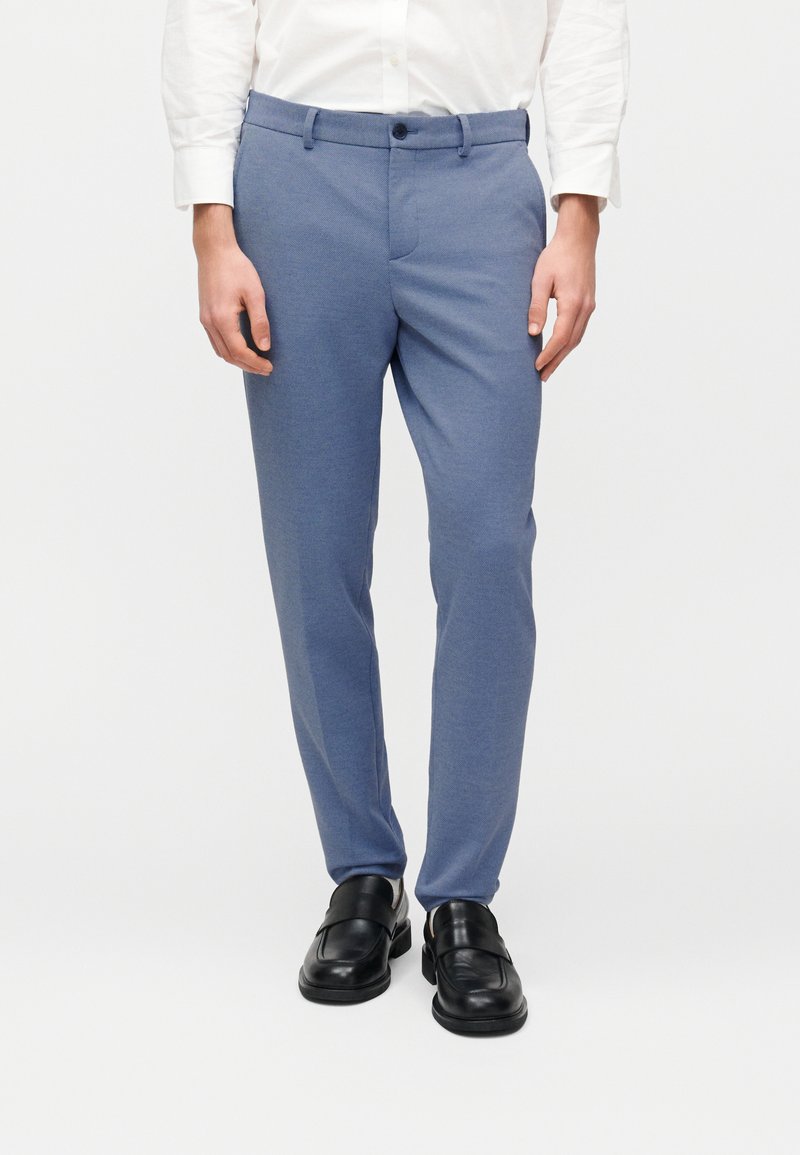 Μπλε παντελόνια ραμμένα από υφασμάτινη υφή, με εφαρμοστή σχεδίαση, πλαϊνές τσέπες και κλείσιμο με κουμπί. Φορεμένα με μαύρα δερμάτινα παπούτσια.