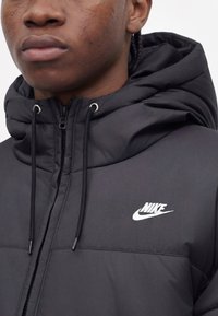 Νεαρό άτομο που φοράει μαύρο μπουφάν Nike με κουκούλα και κορδόνια, εμφανίζοντας το πάνω μέρος του στήθους και το κάτω μέρος του προσώπου.