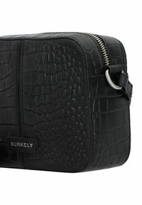 Sac en cuir noir à motif crocodile de forme rectangulaire, avec une fermeture éclair en argent et une étiquette noire de marque sur le côté.