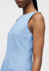 Γυναίκα φορεί ένα αμάνικο, εφαρμοστό, ανοιχτό μπλε φόρεμα με ψηλό λαιμόκοψη, στέκεται μπροστά σε απλό φόντο.