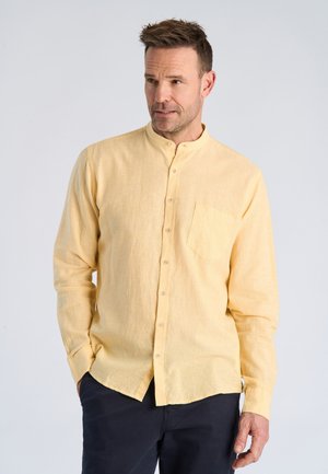 Mann trägt ein hellgelbes Hemd mit Stehkragen und dunkle Hosen, schaut leicht zur Seite und hat die Hände in den Taschen.