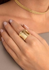 Goldfarbener Metallring mit einem wellenförmigen Design, das eine polierte Oberfläche und ein breites Band aufweist. Trägt an einer Hand mit neutralem Nagellack.