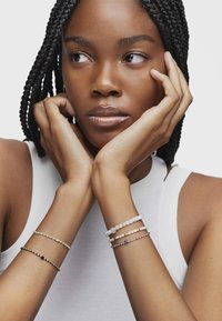 Quattro braccialetti: uno dorato con accenti neri, uno di perline bianche, uno verde e rosa, e uno con dettagli viola. Indossati su un polso.