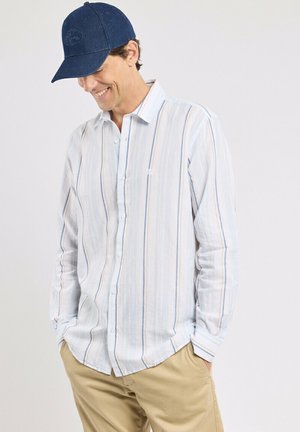 Homme portant une casquette de baseball bleue, une chemise claire à manches longues rayée verticalement, et un pantalon beige, souriant et regardant vers le bas.
