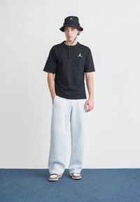 Nuori mies, jolla on musta bucket-hattu, musta t-paita valkoisella logolla, vaaleansiniset leveät housut ja harmaat lenkkarit, seisomassa sinisellä matolla.