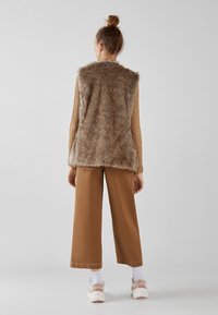 Vesta z umělé kožešiny v hnědé barvě s texturovaným povrchem, doplněná širokými hnědými kalhotami a bílými ponožkami, k tomu tenisky s růžovými akcenty.