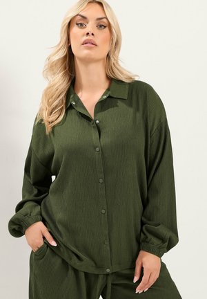 Vrouw met blond haar, gekleed in een loszittend donkergroen hemd met knopen en bijpassende broek, poseert met één hand in de zak.