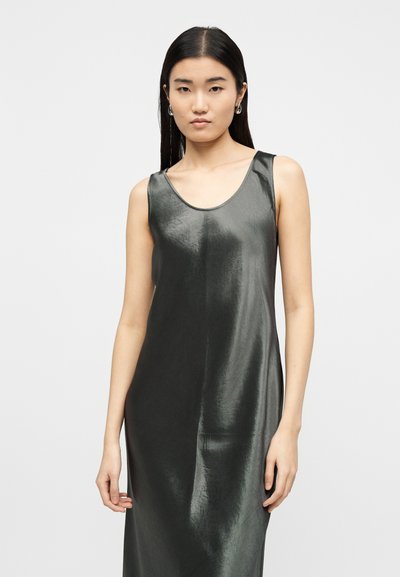 Νεαρή γυναίκα με μακριά σκούρα μαλλιά, φορώντας αστραφτερό μαύρο φόρεμα χωρίς μανίκια και ασημένια σκουλαρίκια κρίκους, μπροστά σε απλό φόντο.