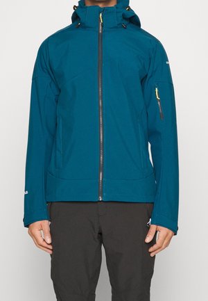 Veste à capuche bleu sarcelle avec fermeture éclair à l'avant et poche zippée sur la manche gauche portée avec un pantalon noir. Mains détendues le long du corps.