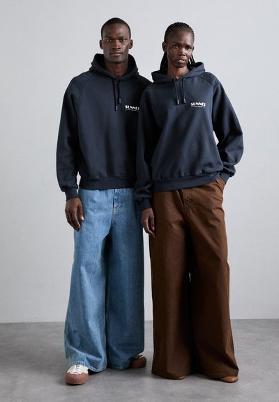 Des sweats à capuche bleu marine avec cordons de serrage, associés à un denim bleu à jambes larges et à des pantalons marron. Les vêtements présentent un logo discret sur les sweats à capuche.