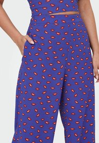 Pantalones de pata de elefante morados con un patrón repetido de labios rojos, que presentan una textura suave y bolsillos, hechos de una tela ligera y suave.