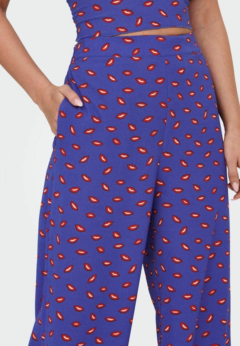 Pantalones de pata de elefante morados con un patrón repetido de labios rojos, que presentan una textura suave y bolsillos, hechos de una tela ligera y suave.